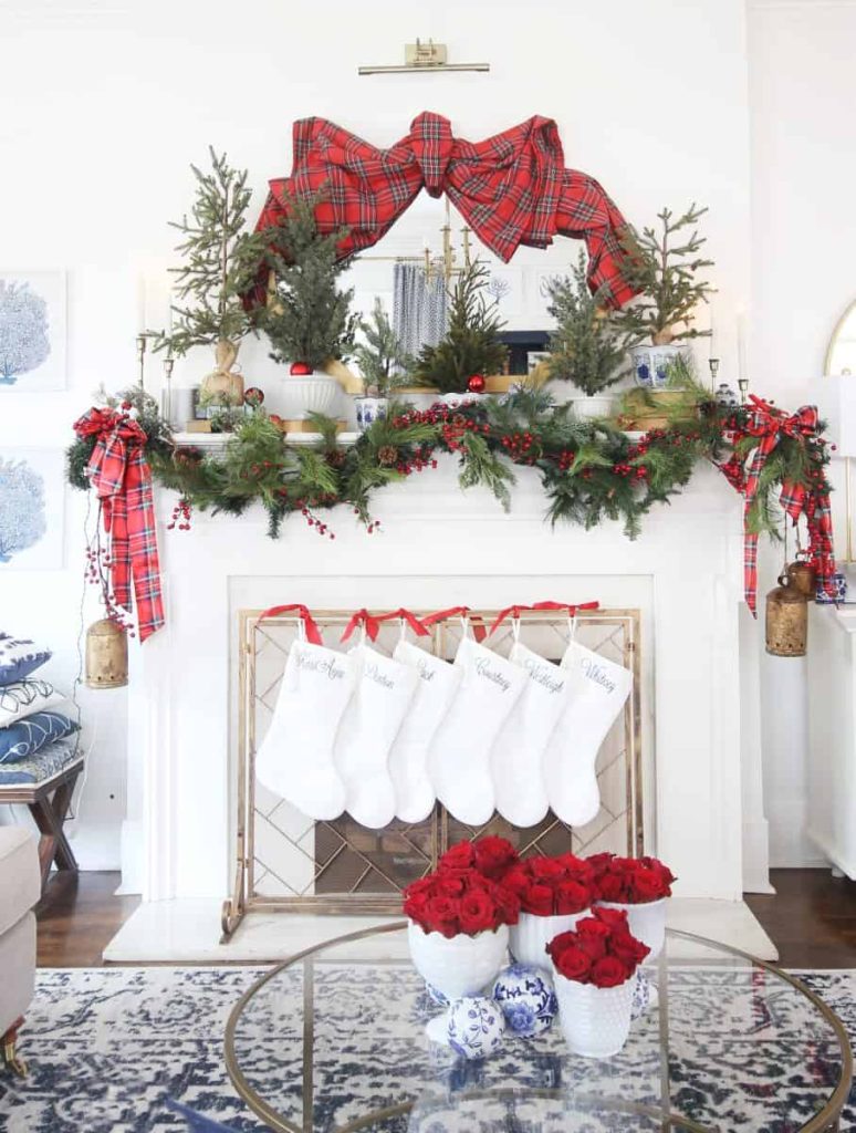 A white fireplace decorated with greenery, red plaid ribbon, and stockings. A large red plaid bow and small Christmas trees sit on the mantel. A bowl of red roses is on the table in front.