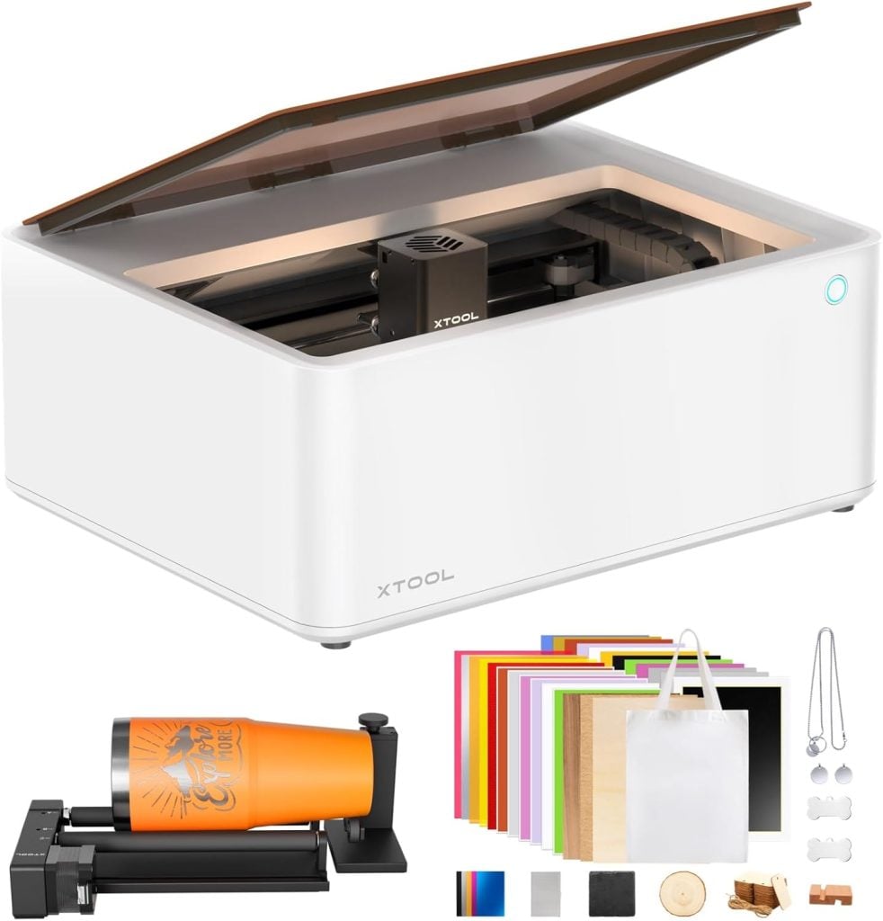 A white xTool laser cutter and engraver machine with an open lid, a rotary attachment, colored material sheets, sample items, and crafting accessories displayed in front.
