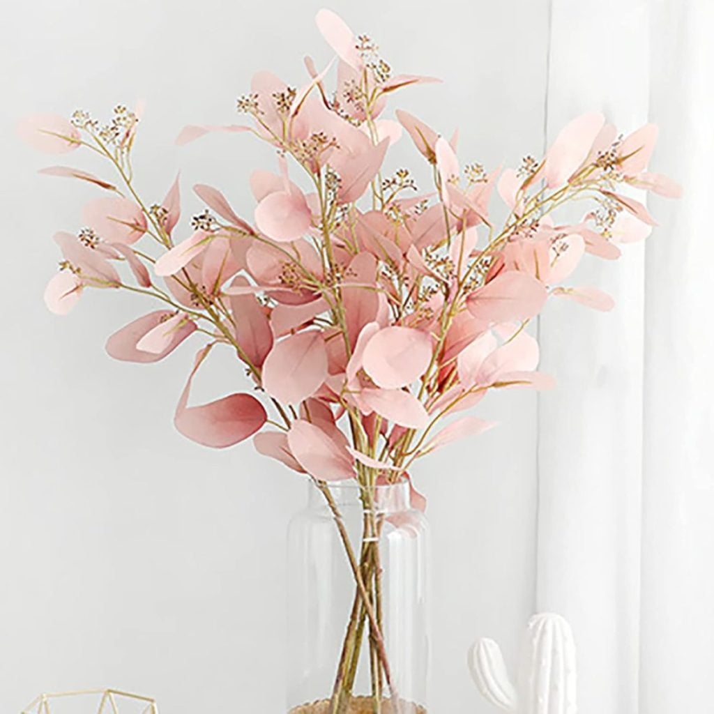 A clear glass vase holds pink eucalyptus stems with round leaves and small white buds, set against a light, neutral background.