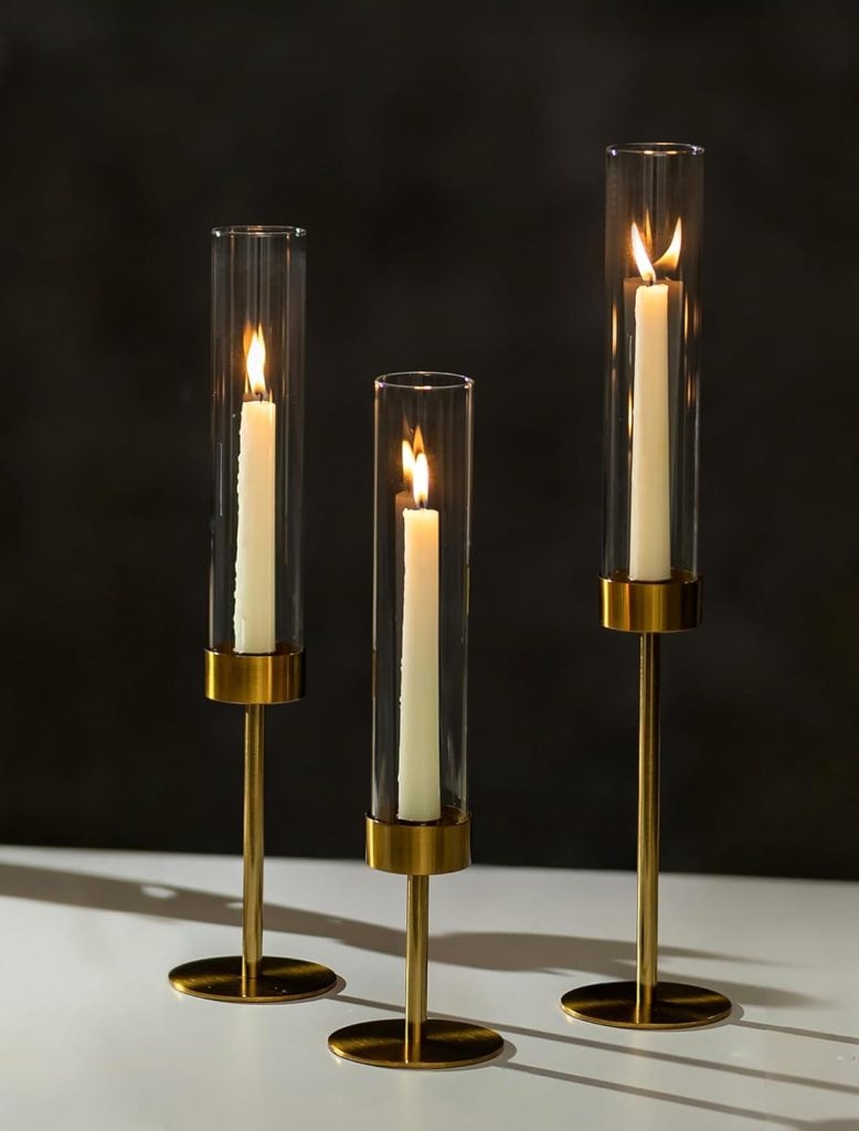 Three lit white candles are placed inside tall, cylindrical glass holders with gold-colored metal bases, arranged on a white surface against a dark background.