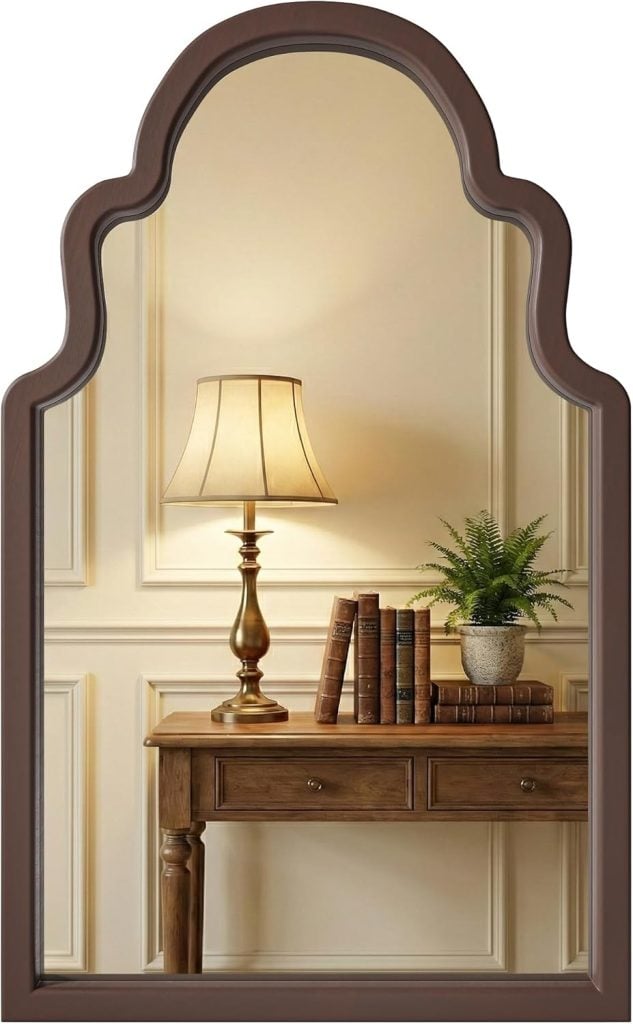 A mirror reflecting a wooden table with stacked books, a brass lamp with a beige shade, and a potted fern against a paneled wall.