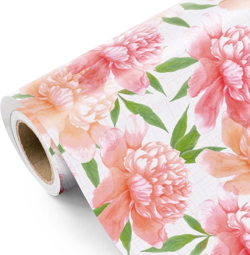 A roll of wallpaper with a pink and peach peony floral pattern and green leaves on a white background.