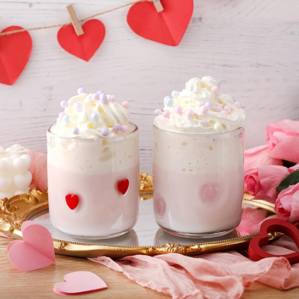 Two glasses of pink milkshakes with whipped cream and pastel sprinkles, decorated with red heart stickers, sit on a gold tray among paper hearts and pink roses.