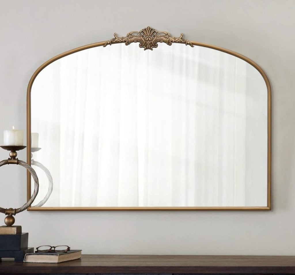 A large, arched wall mirror with a decorative gold frame hangs above a wooden table holding a candle, books, and glasses.