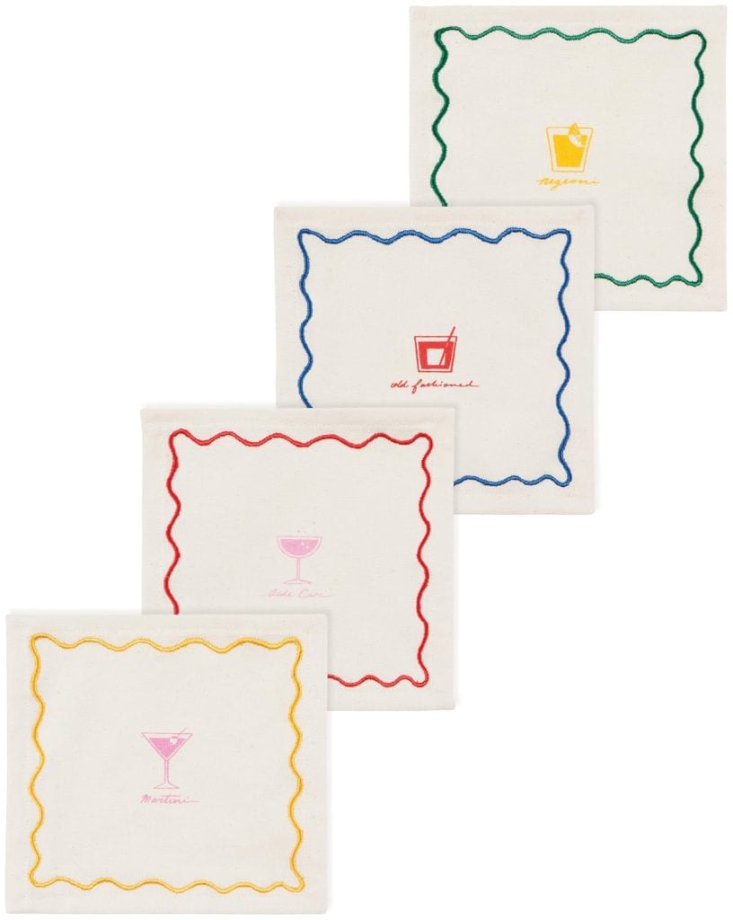 Four white fabric cocktail napkins with colored wavy borders and embroidered images of drinks labeled "Negroni," "Old Fashioned," "Whiskey Sour," and "Martini.