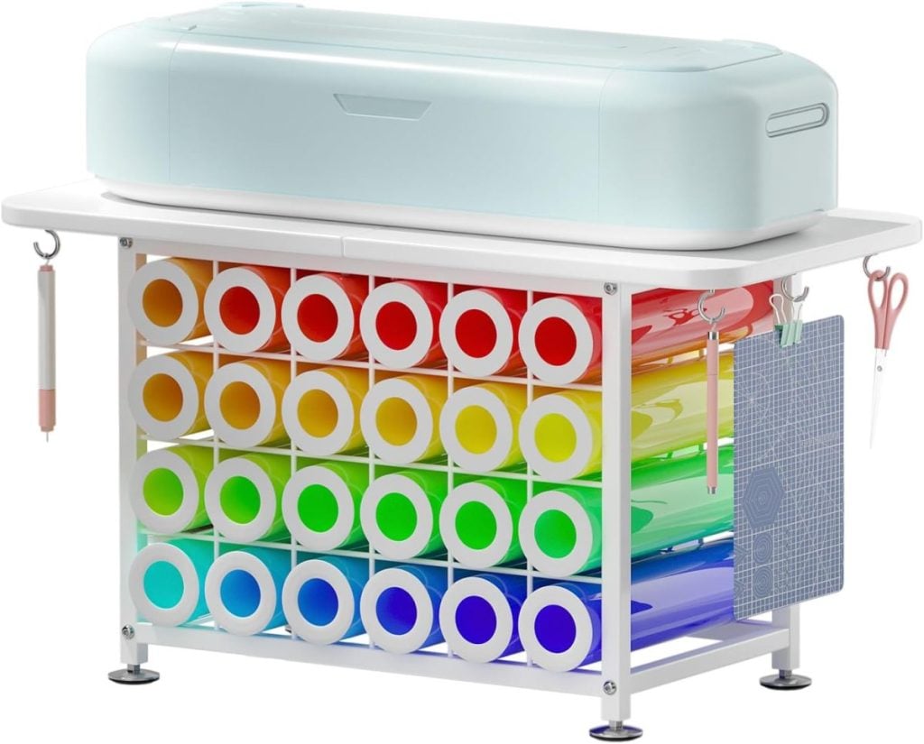 A craft cutting machine sits on a rack holding multiple rolls of colored vinyl arranged in a gradient, with tools and a cutting mat hanging on the sides.