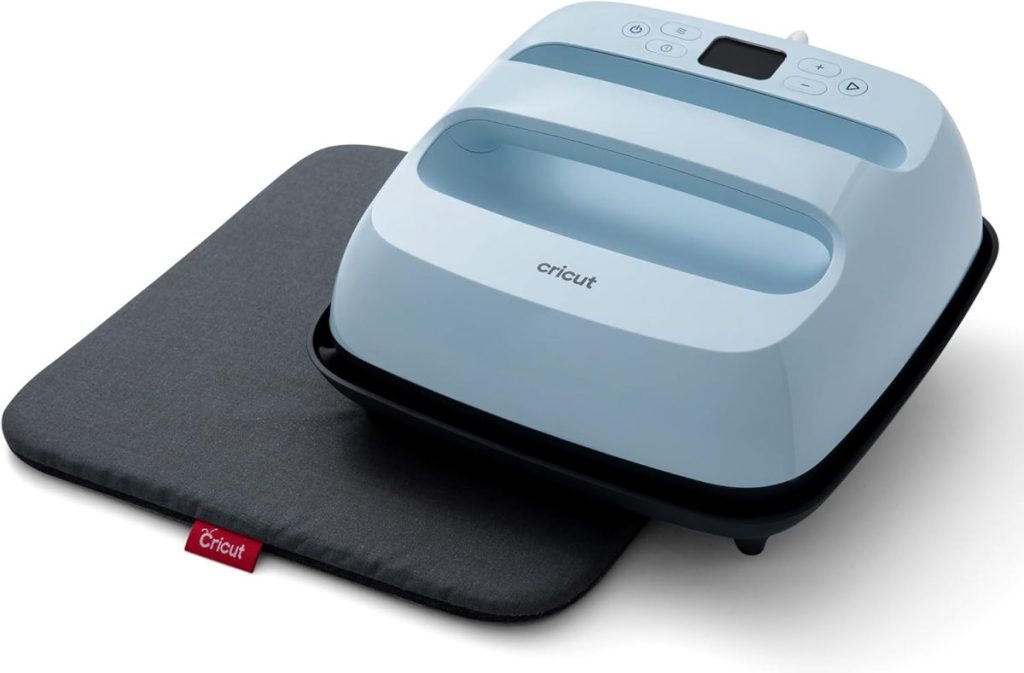 A Cricut heat press in light blue rests on a dark gray heat-resistant pad, with control buttons and a display visible on the device.
