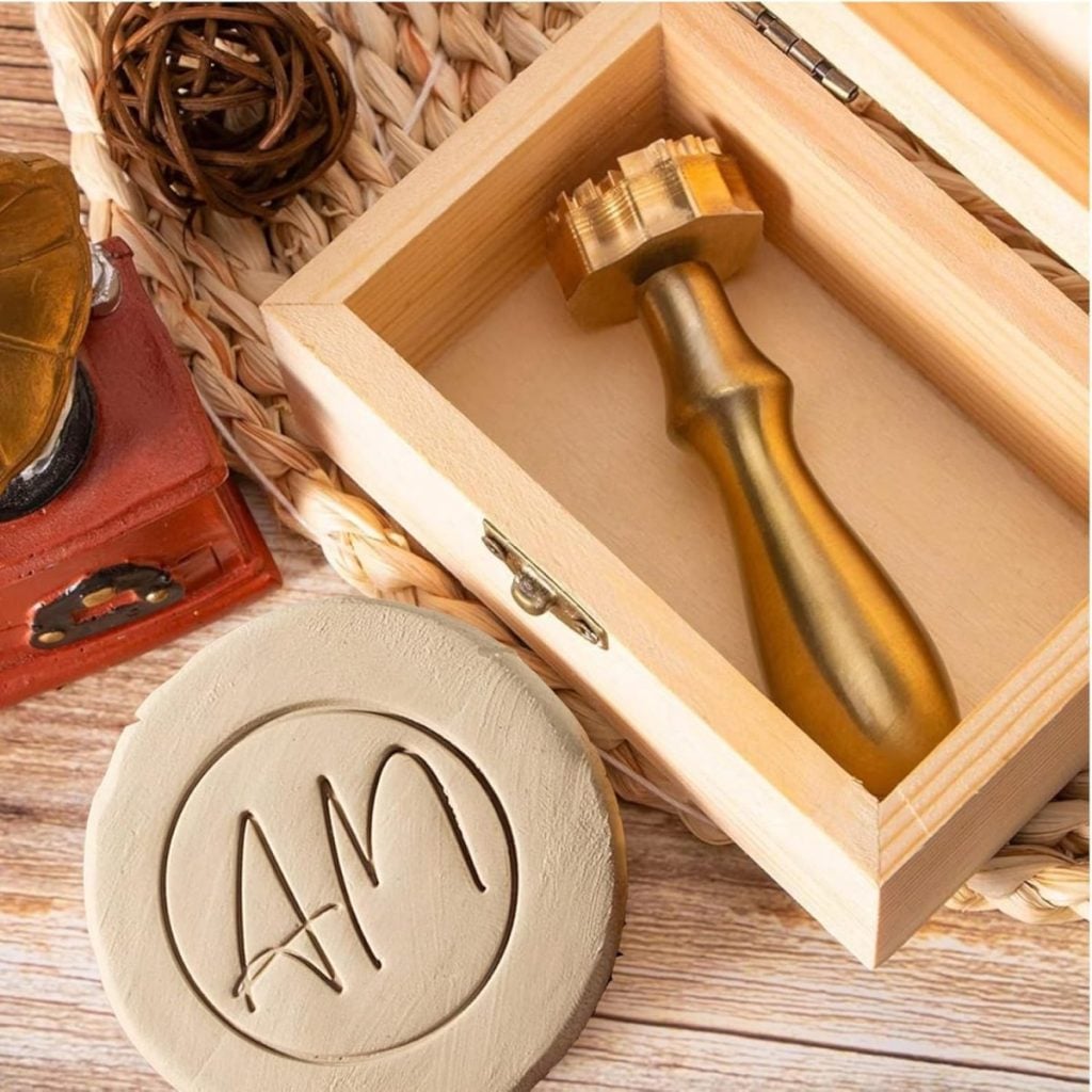A gold wax seal stamp with the initials "AM" sits in an open wooden box, next to a stamped clay disc displaying the same initials.