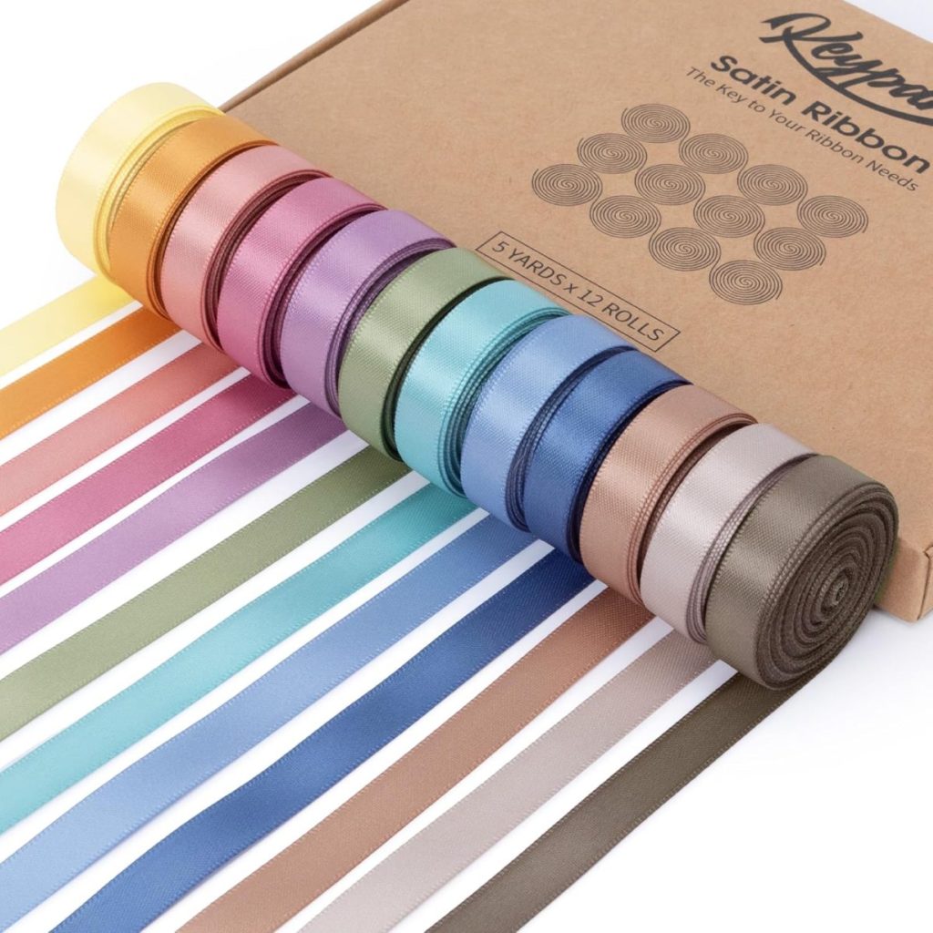Twelve rolls of satin ribbon in assorted colors are displayed next to a brown Kleymax box labeled "Satin Ribbon." Each ribbon is partially unrolled in a straight line.