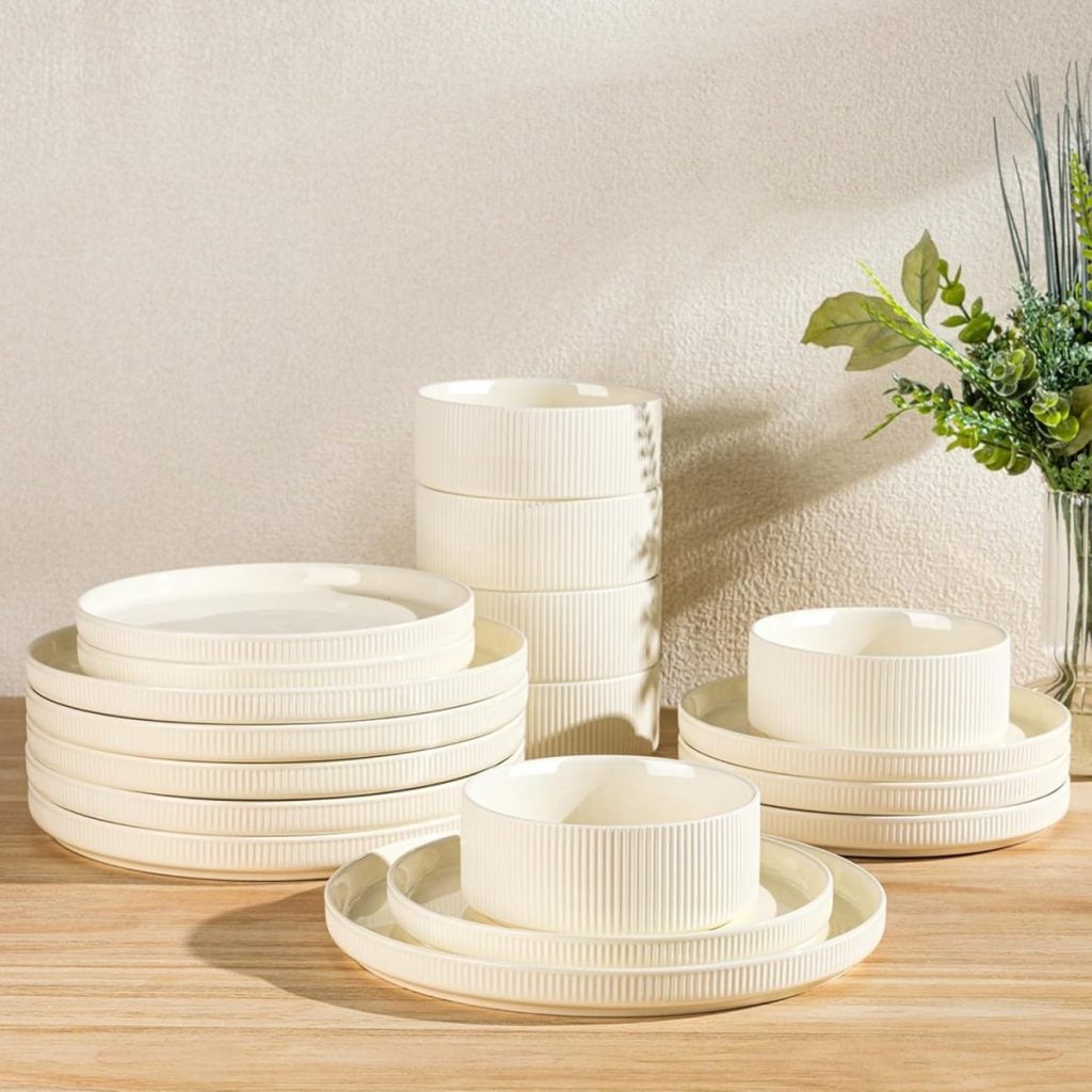 A set of cream-colored ceramic dishes, including plates and bowls with ribbed texture, arranged on a wooden surface next to a green floral arrangement.
