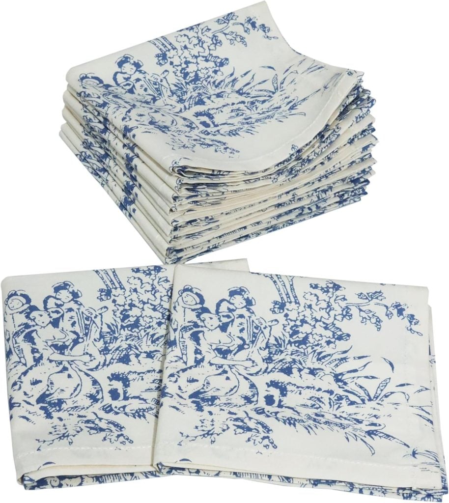 A stack of folded white cloth napkins with a blue pastoral pattern, and two additional napkins laid flat in front.