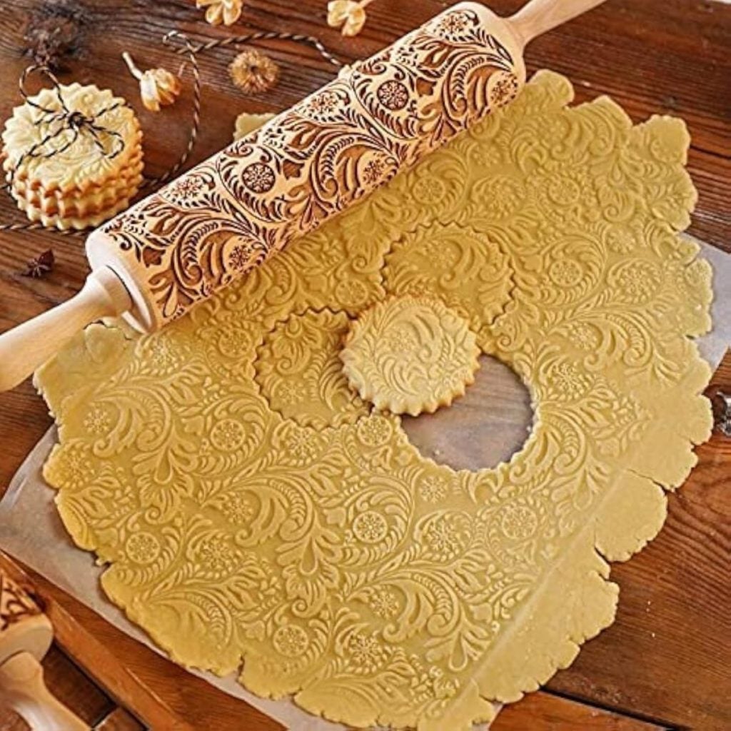 Embossed rolling pin creates intricate floral patterns on rolled-out cookie dough; a round cookie cutter has been used to cut one patterned cookie.