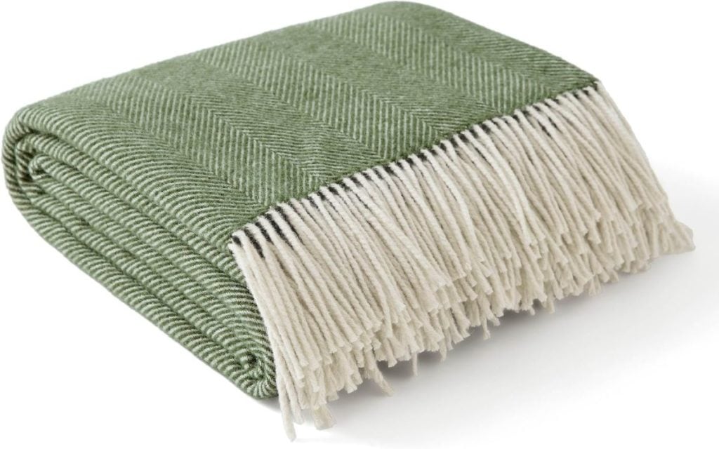 A folded green herringbone-patterned blanket with off-white fringed edges, placed on a white surface.