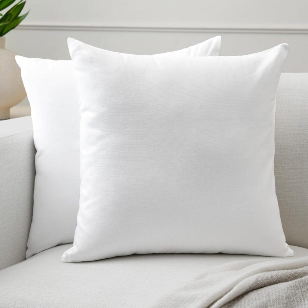 Two plain white square pillows on a light-colored sofa with a light blanket and a bit of green plant in the background.