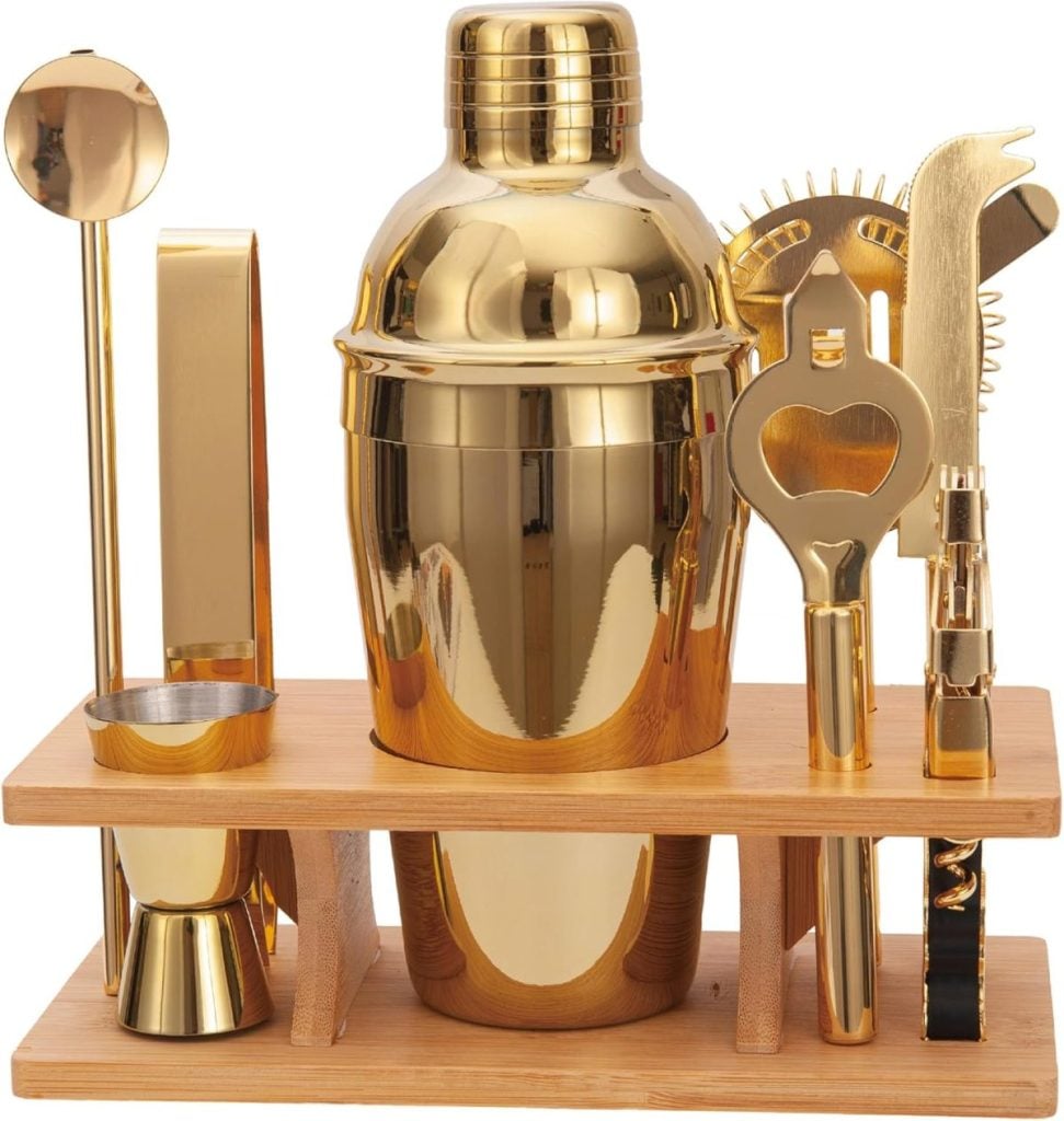 A gold-colored cocktail shaker set with various bar tools, including a jigger, strainer, opener, tongs, and stirrer, organized on a wooden stand.