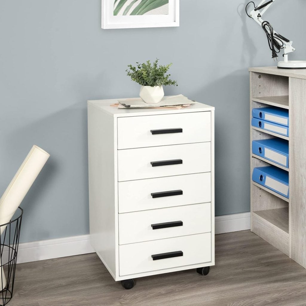 A white five-drawer storage cabinet on wheels is placed against a light gray wall, next to a desk with blue binders and a lamp. A potted plant sits on top of the cabinet.