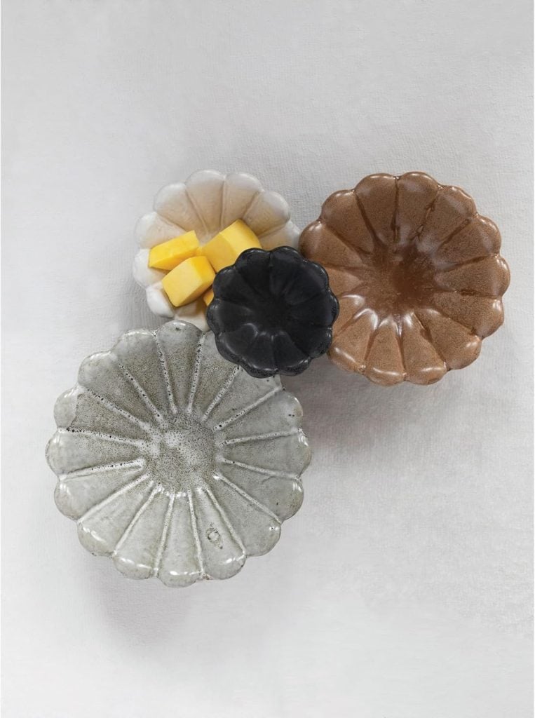 Four flower-shaped ceramic dishes in different colors, one containing four cubes of butter, arranged on a light textured surface.