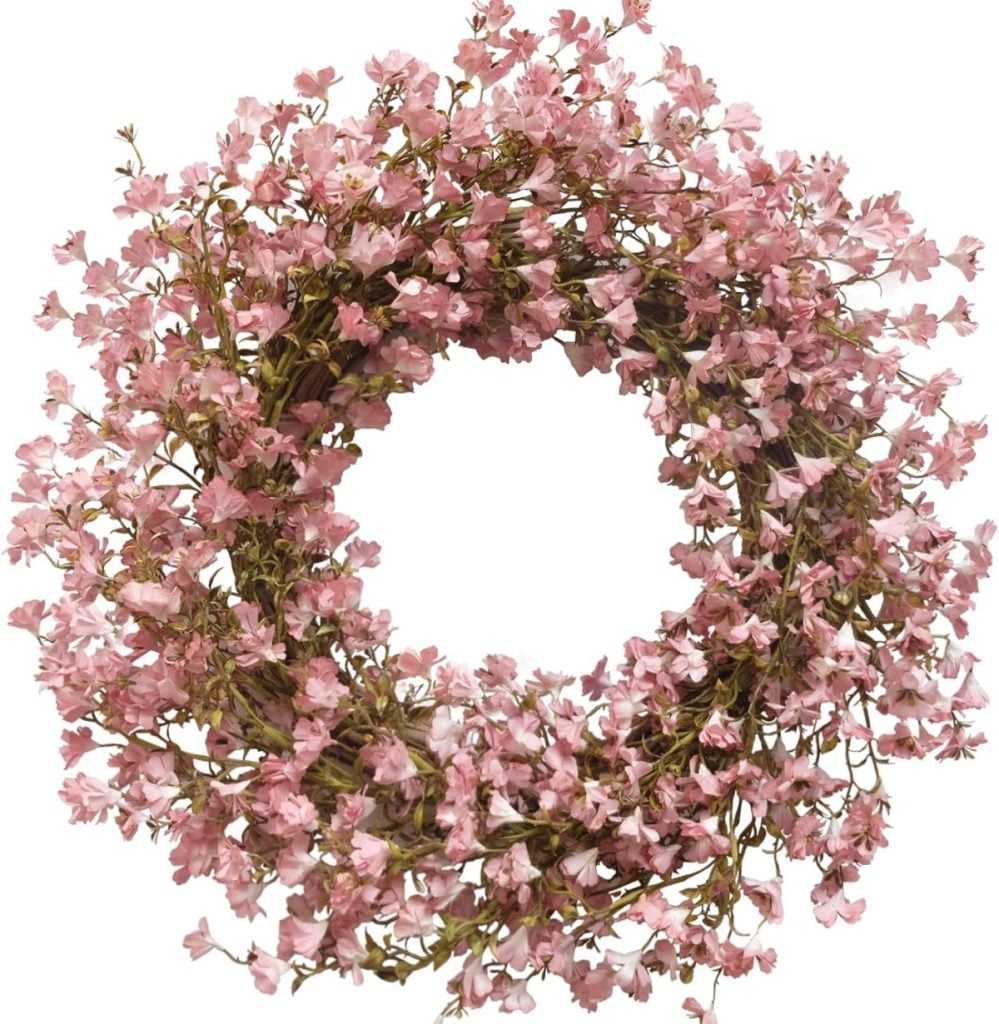 A round wreath made of small, pink artificial flowers with green stems, arranged densely in a circular shape on a white background.