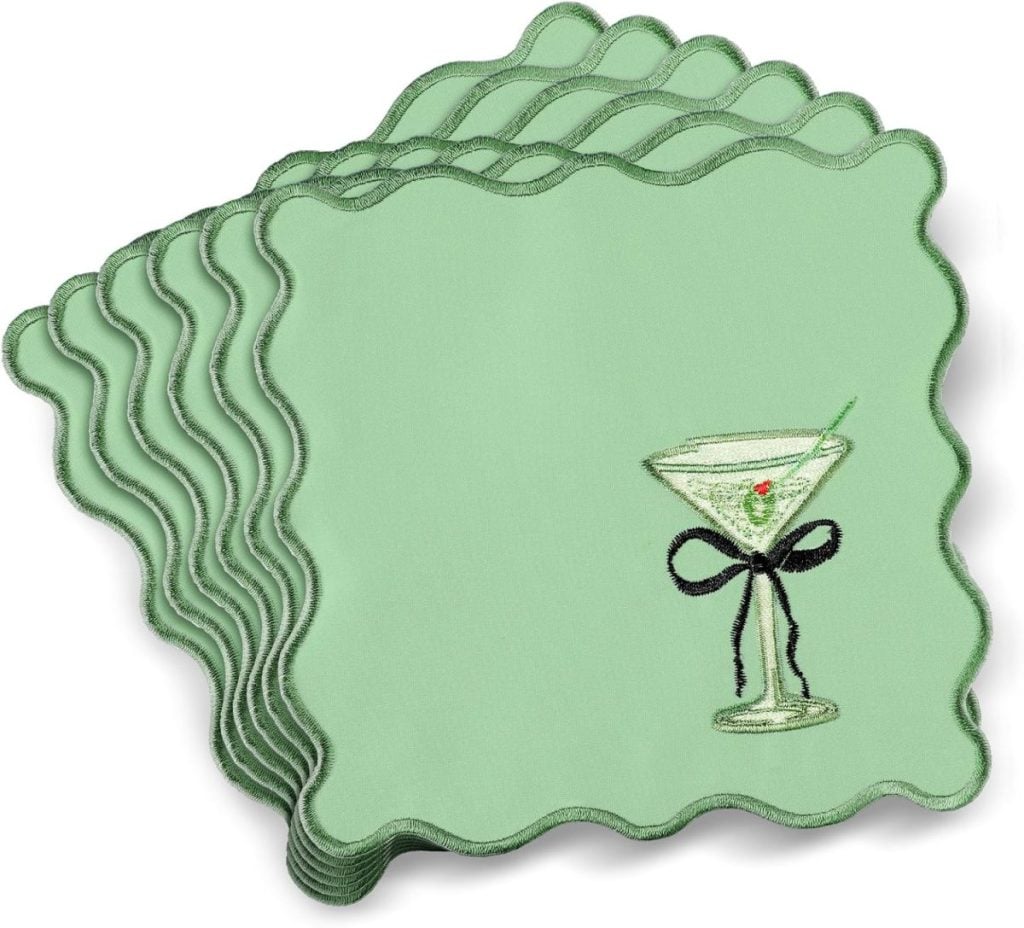A stack of six green scalloped-edge cocktail napkins, each embroidered with a martini glass, olive, and a black bow.