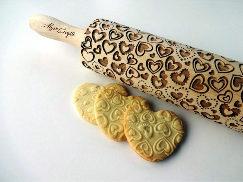 A wooden rolling pin engraved with heart shapes lies next to three heart-shaped cookies embossed with the same heart pattern.
