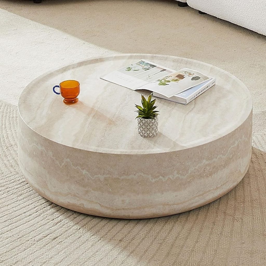 Round beige stone coffee table with a small potted plant, an orange glass cup, and an open magazine on top, placed on a textured light rug.
