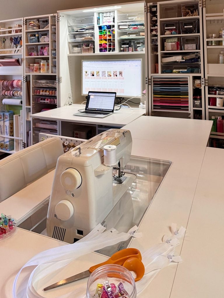 Organized sewing room with a sewing machine on a white table, open laptop, scissors, zippers, and various sewing supplies stored neatly on shelves in the background.