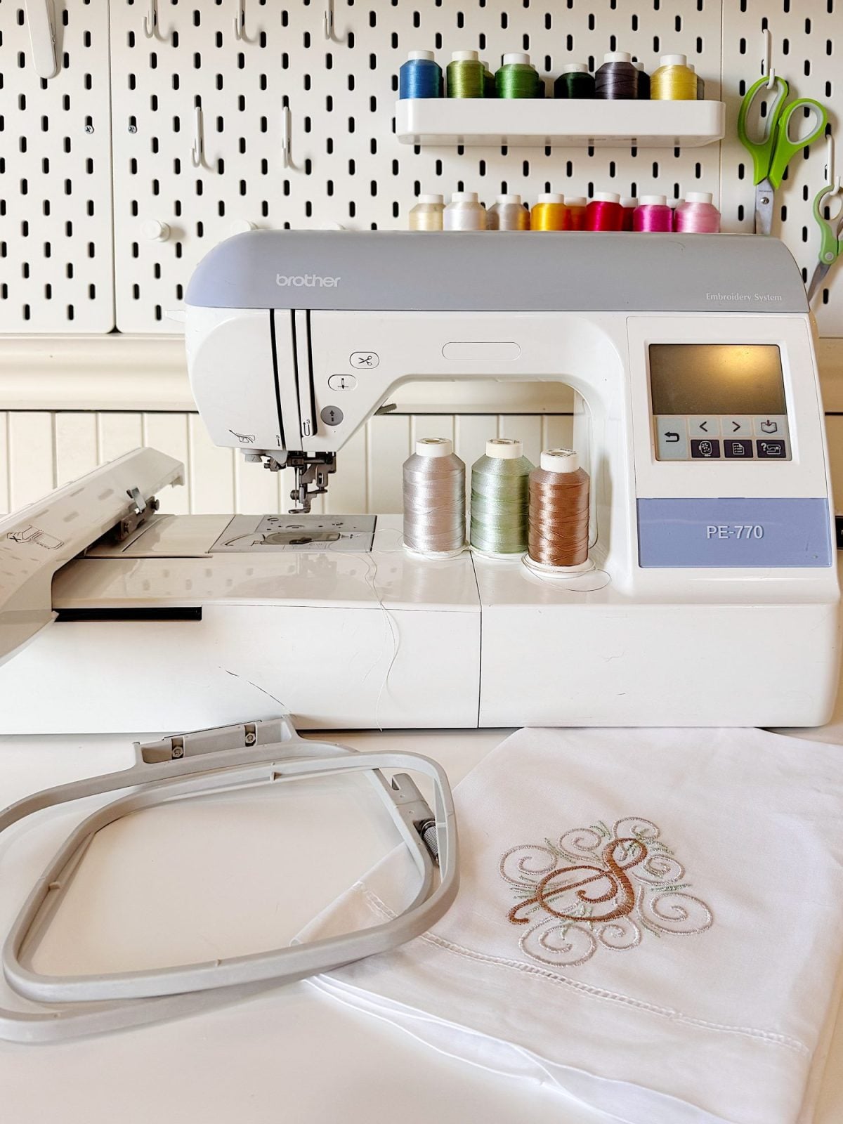 A Brother PE770 embroidery machine with thread spools, green scissors, and colored thread on shelves, with an embroidery hoop and stitched fabric in the foreground.