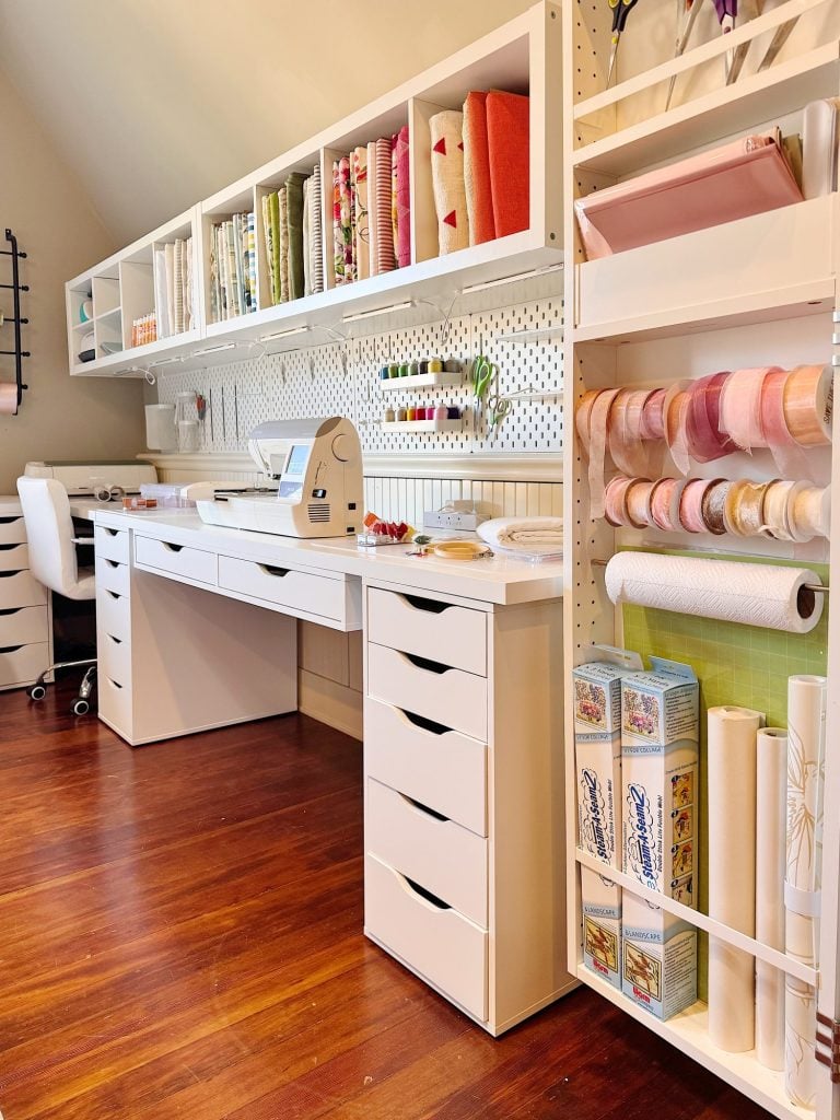 A well-organized craft room with white shelving, drawers, and a pegboard wall, storing fabric, ribbons, craft tools, and a cutting machine on a tidy wooden floor.