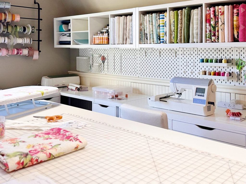 A well-organized sewing room with a sewing machine, ironing board, fabric rolls, spools of thread, and ribbons neatly arranged on shelves and work surfaces.