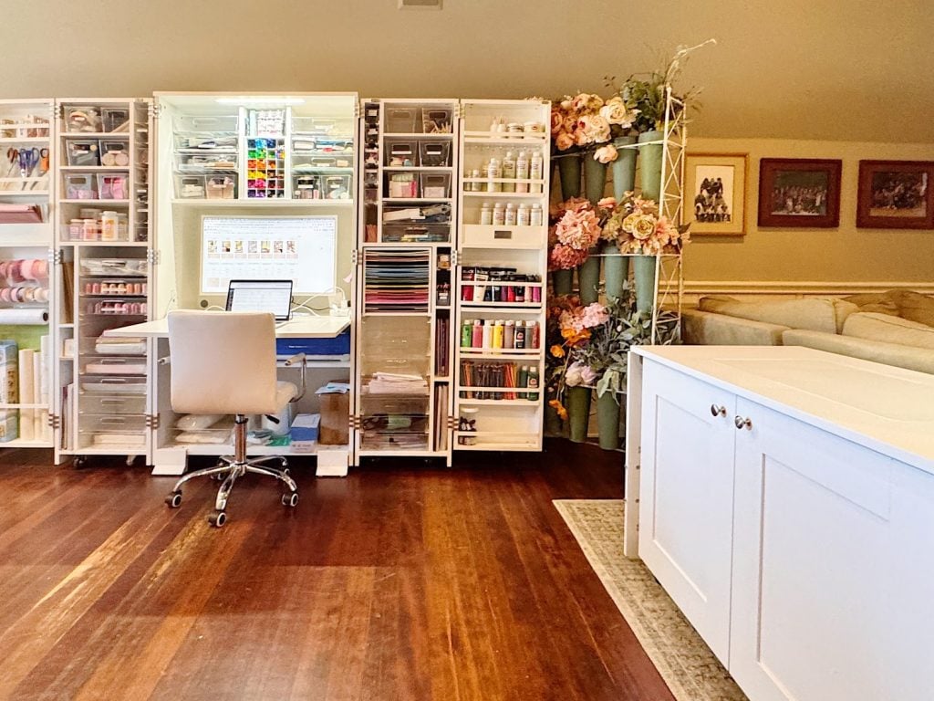 A neatly organized craft room with open storage cabinets filled with art supplies, a white desk and chair, and flower arrangements on display.