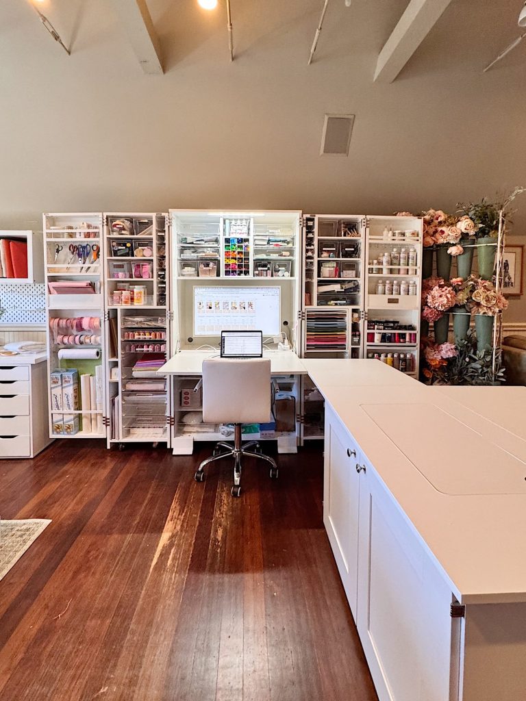 A craft room with an organized white desk, storage cabinets filled with supplies, a rolling chair, and shelves holding art materials and decorative flowers.