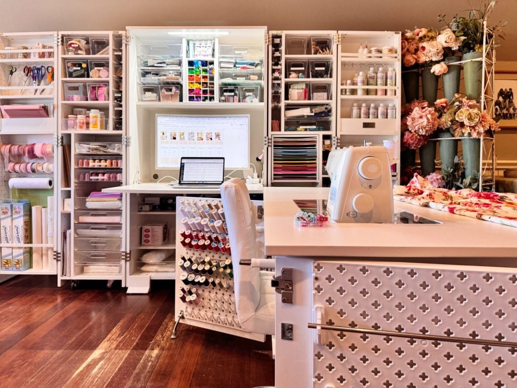 A well-organized craft room featuring open shelves with supplies, a sewing machine on a desk, spools of thread, and rolls of ribbon and fabric.