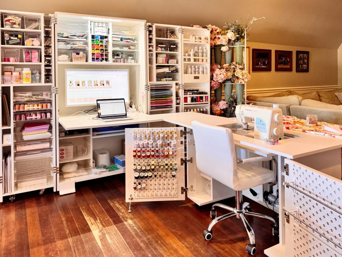 A well-organized craft room with storage cabinets, a computer, sewing machine, thread spools, and craft supplies arranged neatly on and around white desks and a white chair.
