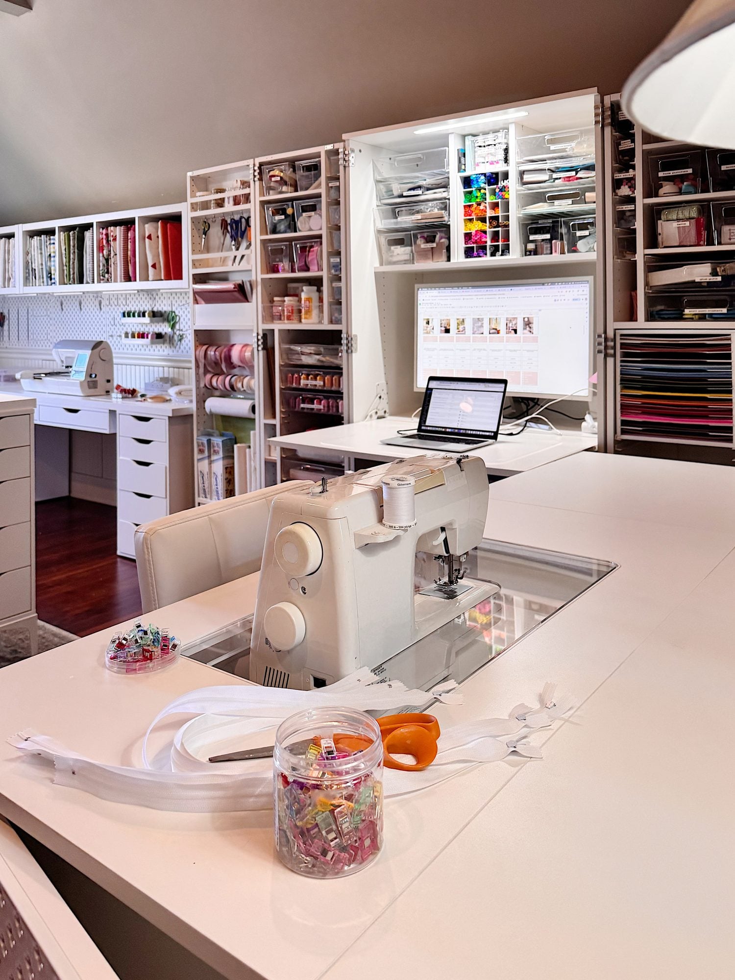 A sewing workspace with a sewing machine, zipper, scissors, and clips on a white table; shelves with craft supplies and a laptop are in the background.
