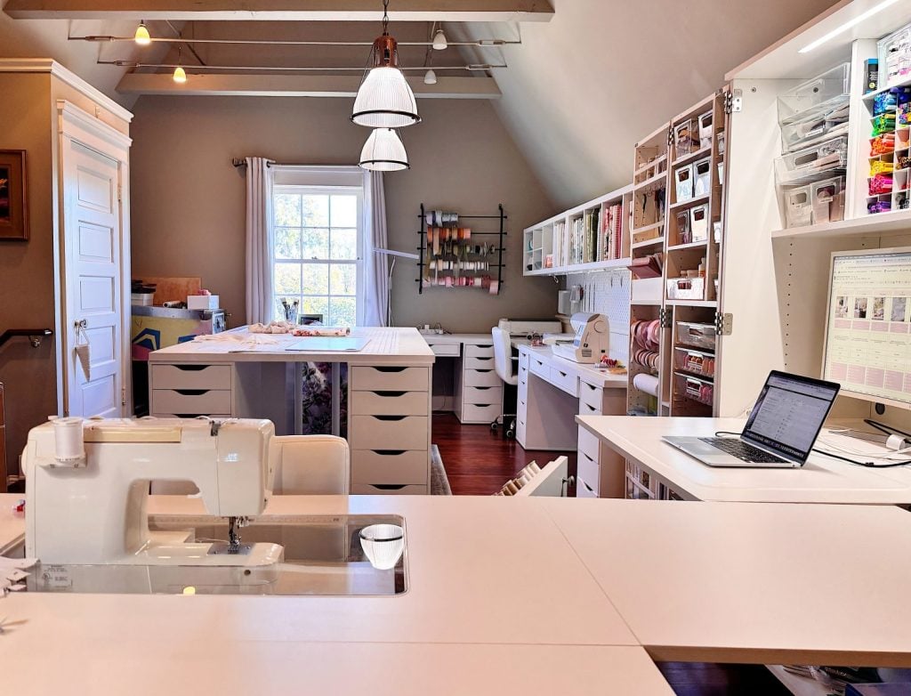 A well-organized craft room with a sewing machine, laptop, shelves of supplies, large worktables, and natural light from a window.
