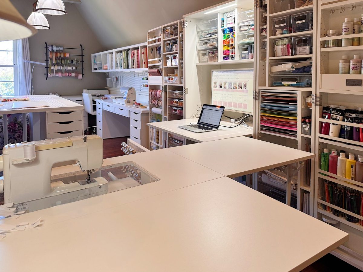 A well-organized craft room with shelves of supplies, a sewing machine, a laptop on a desk, and ample workspace under bright lighting.