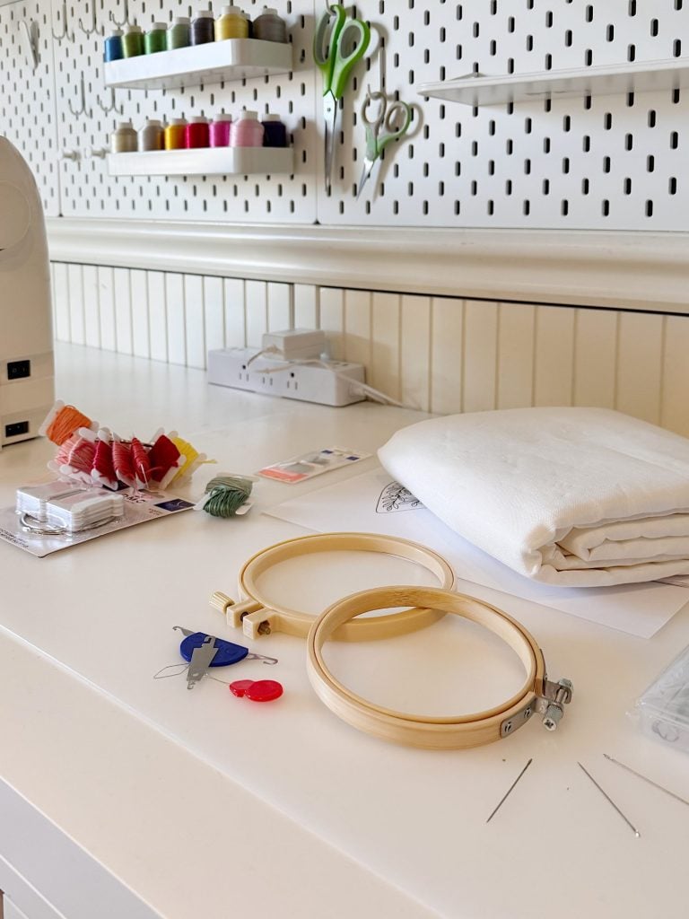 Embroidery supplies on a white table, including fabric, embroidery hoops, threads, needles, and sewing tools; spools of thread and scissors hang on a pegboard in the background.