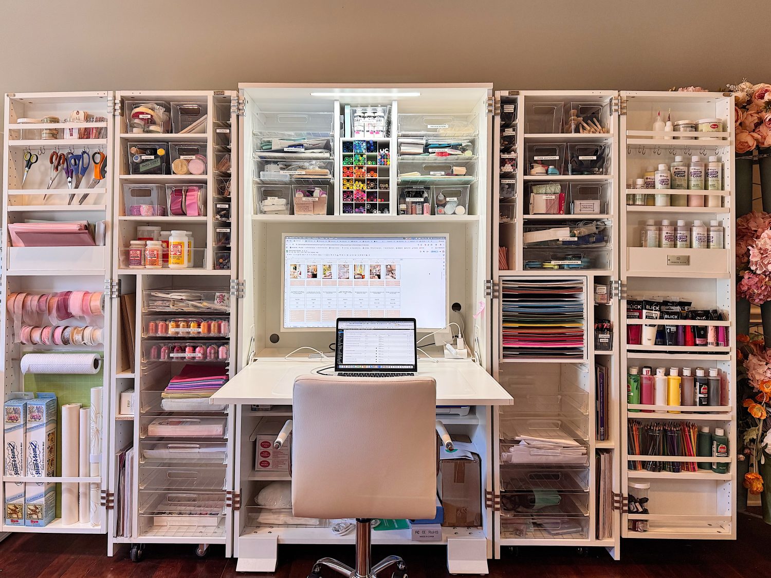 Well-organized craft workspace with shelves of supplies, a central computer monitor, laptop on desk, and various crafting materials neatly arranged around a white chair and workstation.