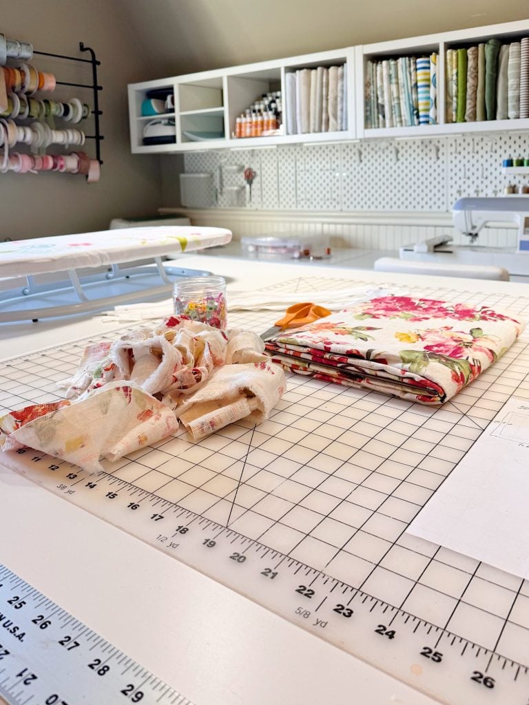 A sewing workspace with fabric, a ruler, and sewing tools on a cutting mat; shelves with supplies and a sewing machine are in the background.