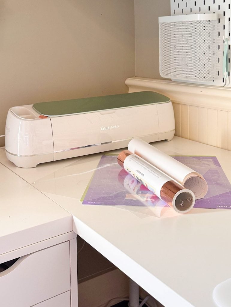 A Cricut Maker cutting machine sits on a white desk next to a purple cutting mat and rolled vinyl sheets in a home craft workspace.