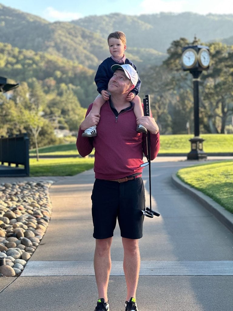A man stands on a golf course path holding a putter, with a young boy sitting on his shoulders. A large clock and mountains are visible in the background.