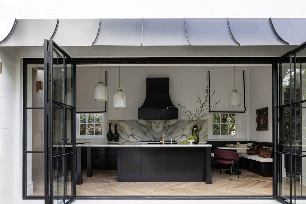 Modern kitchen with black island, marble backsplash, and gold accents, viewed through wide open black-framed glass doors. Pendant lights hang above island; seating nook is on the right.