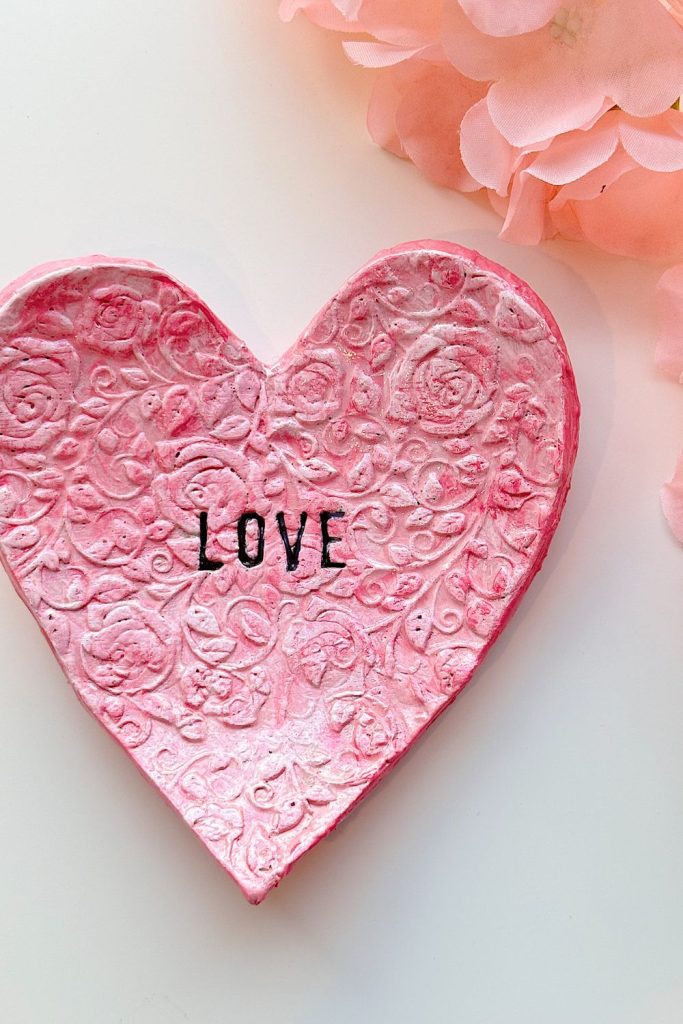 A pink, textured heart-shaped decoration with floral patterns and the word "LOVE" written in black, placed beside light pink flowers on a white surface.