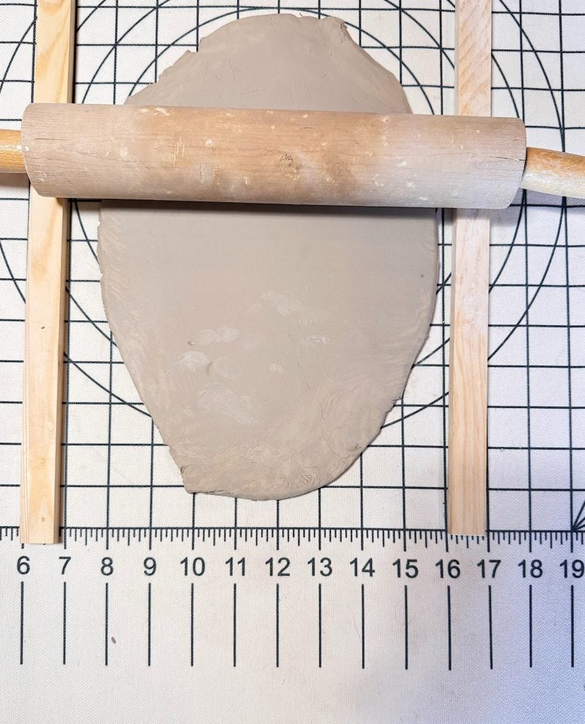 A rolling pin flattens a slab of clay between two wooden slats on a gridded work surface with a ruler at the bottom.