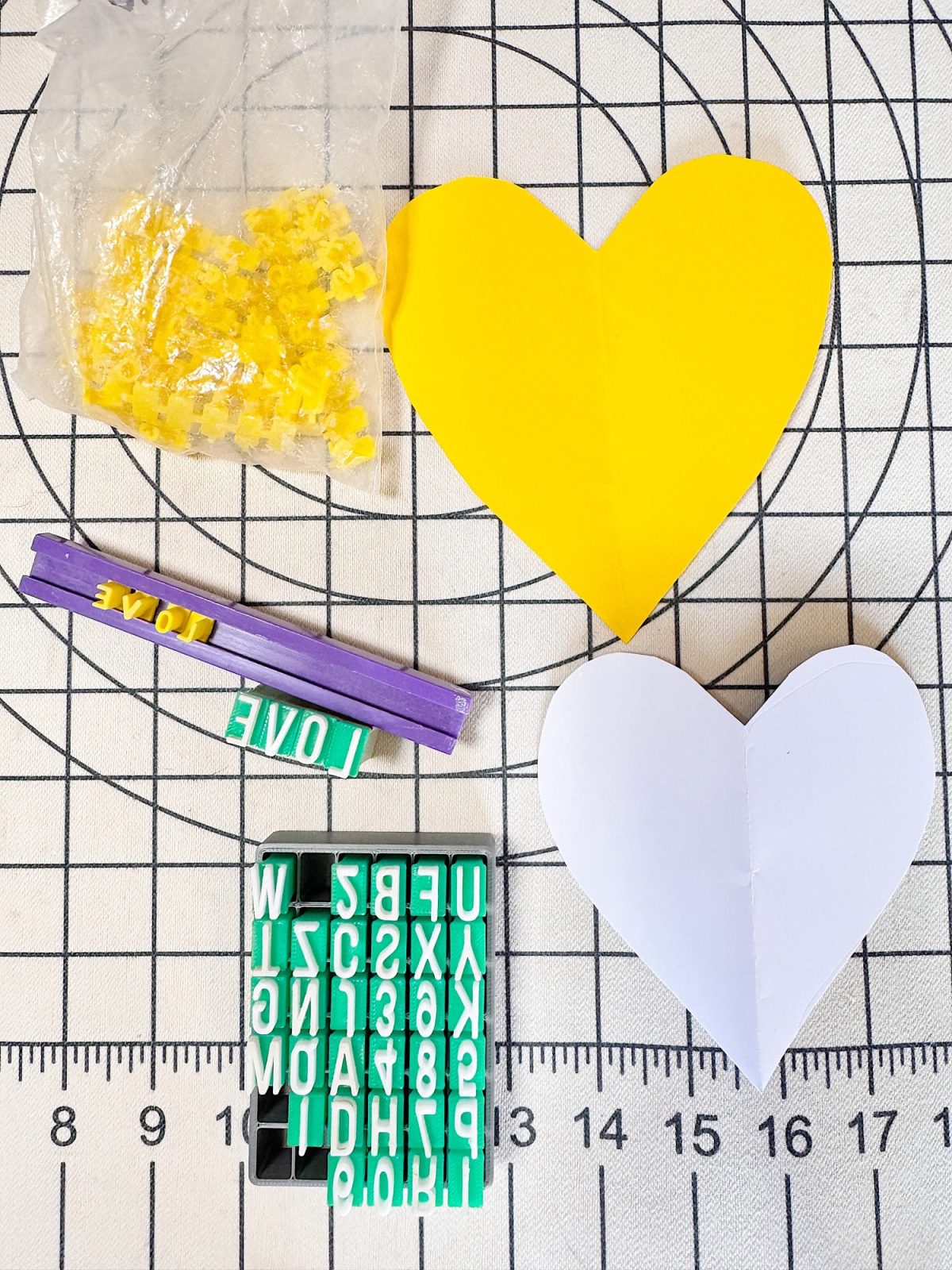 Yellow and white paper hearts, yellow plastic pieces in a bag, a purple tool with "LOVE" stamps, and a green alphabet stamp set on a gridded cutting mat.