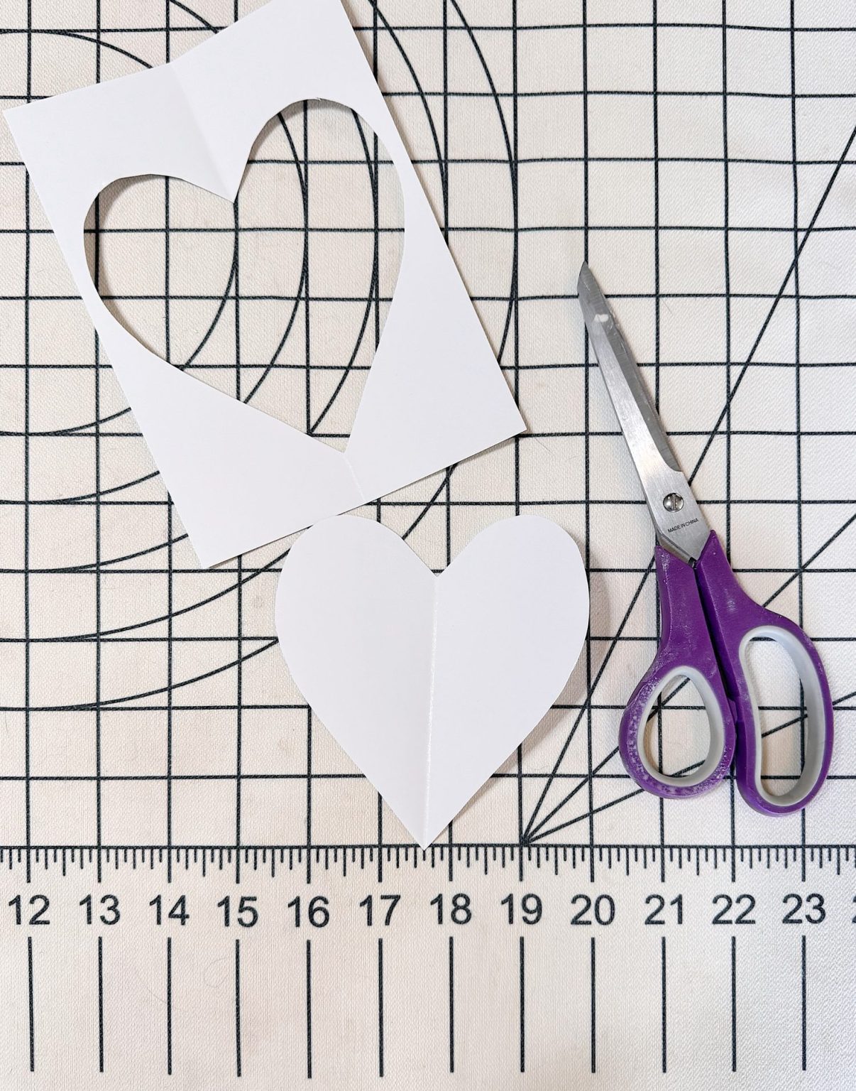 A piece of paper with a heart shape cut out, the cut-out heart, and purple-handled scissors are placed on a gridded cutting mat with measurements.