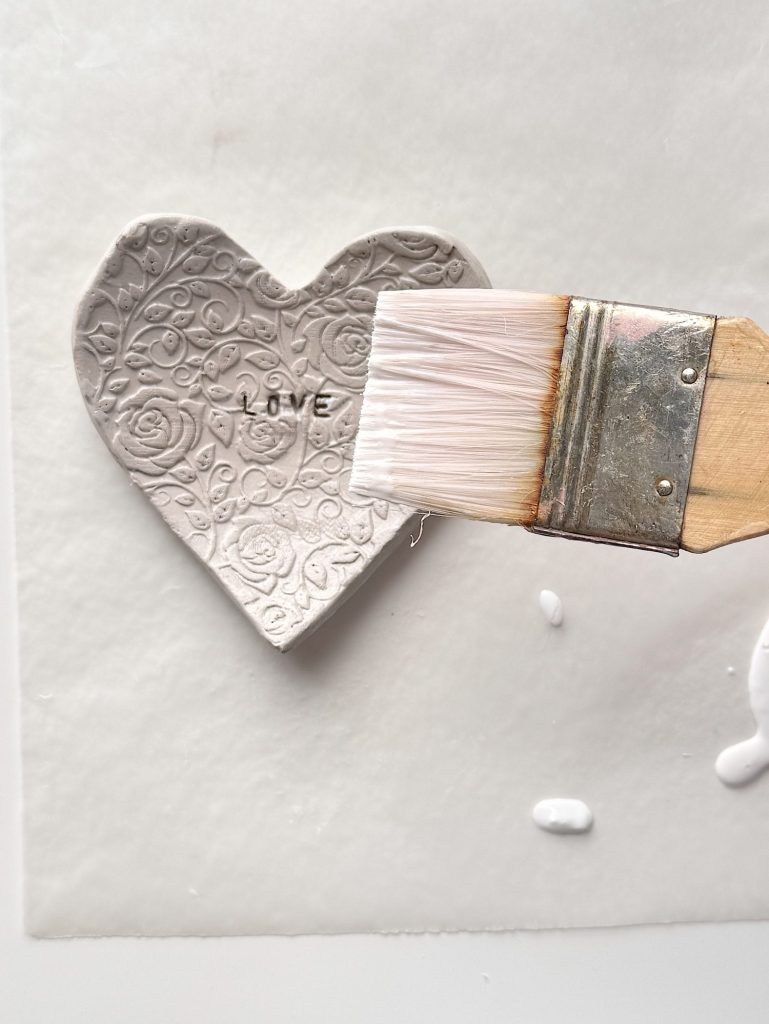A paintbrush applies white glaze to a textured clay heart ornament with the word "LOVE" stamped in the center.