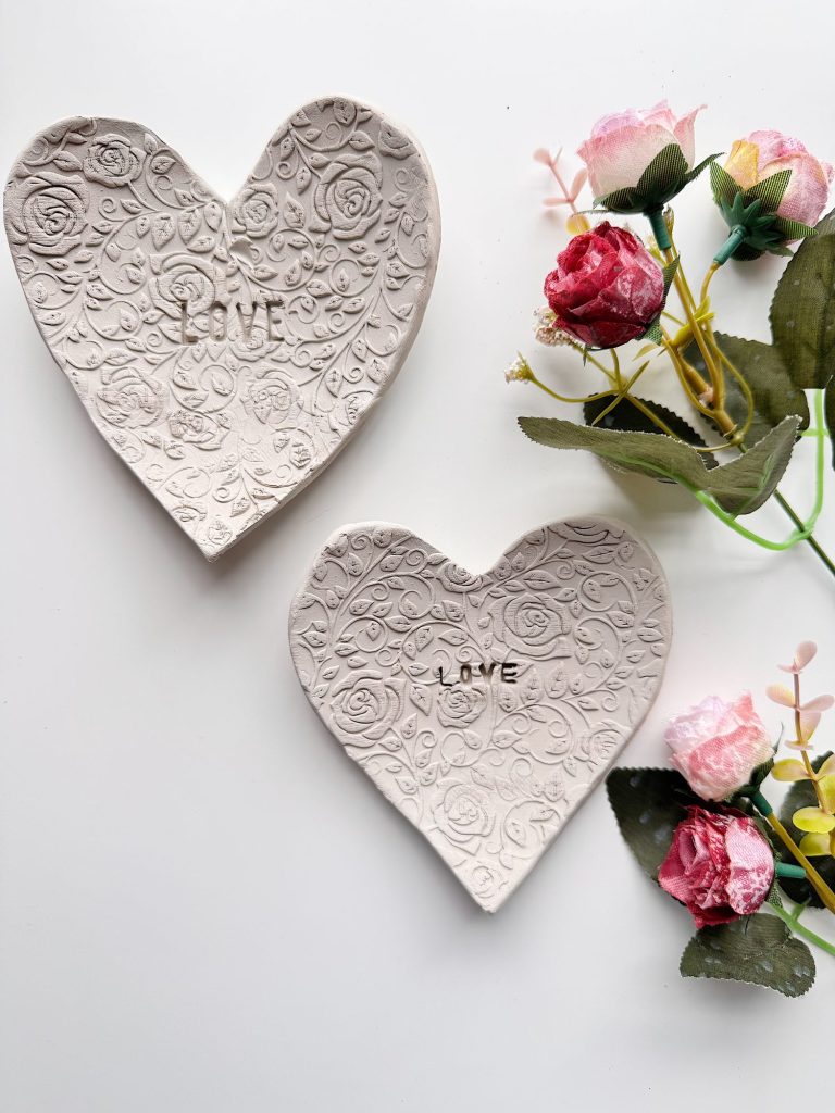 Two heart-shaped ceramic dishes with a floral pattern and the word "LOVE" engraved, placed beside artificial pink and red flowers on a white surface.