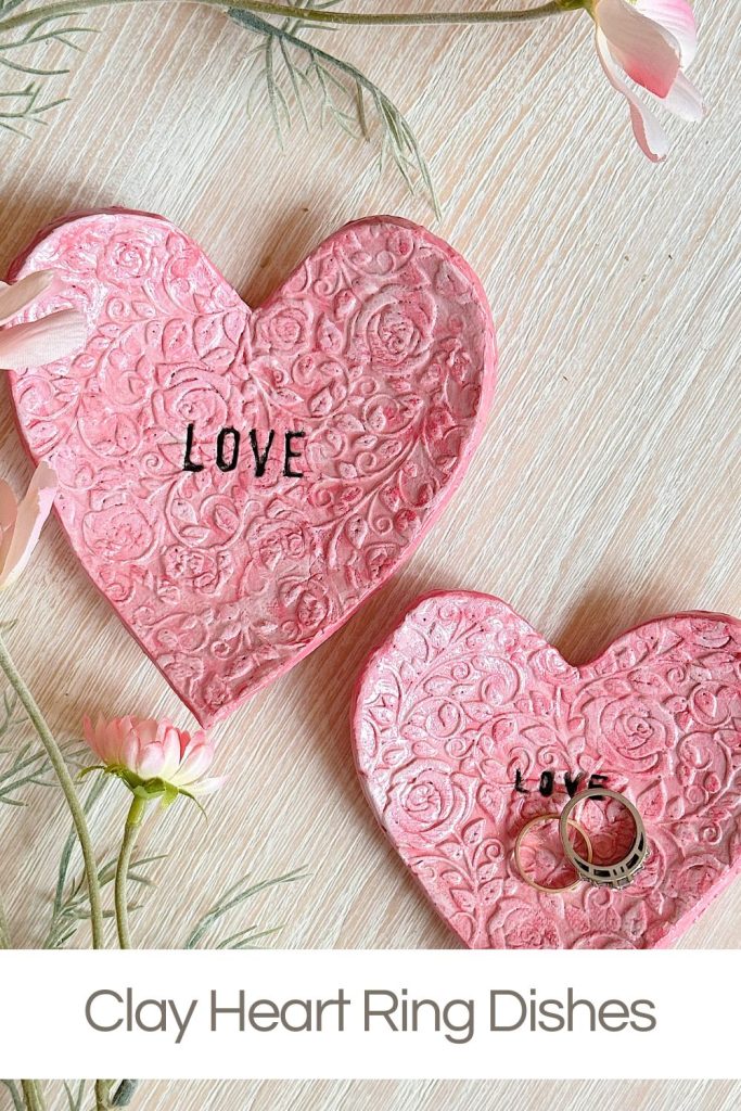 Two pink, heart-shaped clay dishes with embossed floral patterns and the word "LOVE," one holding two rings, on a light wooden surface with flowers.