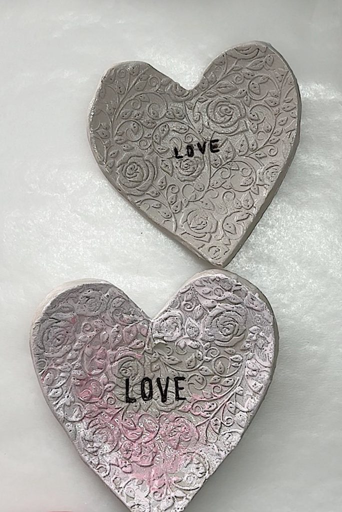 Two heart-shaped clay decorations with embossed floral patterns and the word "LOVE" in black letters, placed on a white surface.